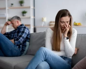 A man and woman appear troubled, seated apart on a couch, each engrossed in thought.