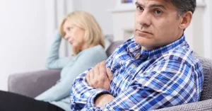 A man and woman sitting on a couch with crossed arms, showing expressions of discontent, facing away from each other, indicative of relationship tension.