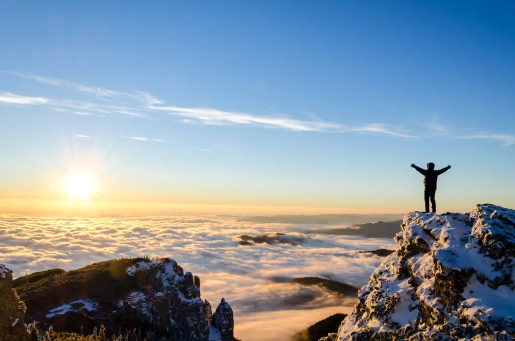 A person stands on a snowy mountain peak with arms raised triumphantly against a backdrop of a sunlit sky above the clouds at sunrise.