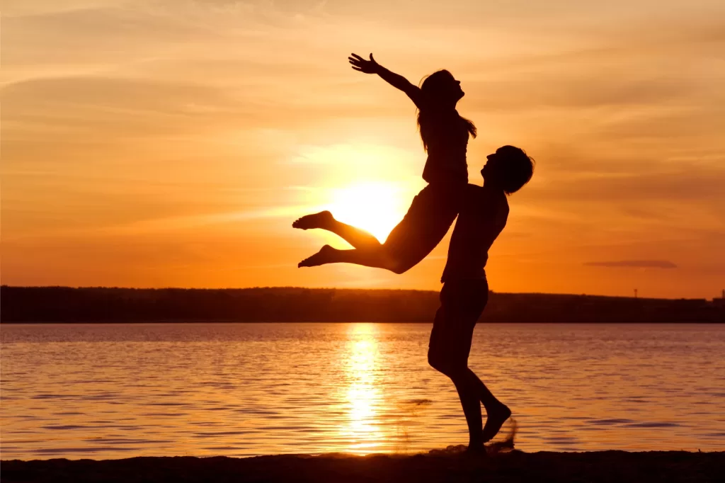 Silhouette of two people on a beach, one lifting the other playfully, against a sunset backdrop reflecting on the water.