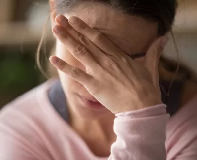 A woman in a pink sweater covers her face with her hand, a gesture of distress or deep thought.