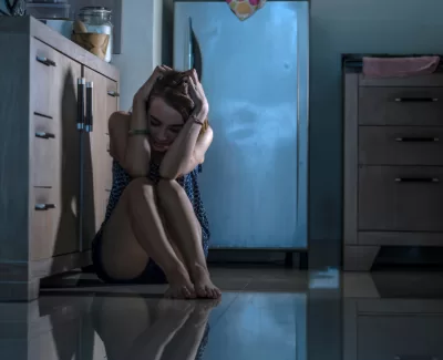 A distressed woman sits on the floor of a kitchen, clutching her head in her hands.